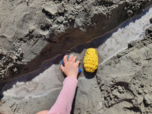 Load image into Gallery viewer, A child&#39;s hand squishing a blue EcoSplat reusable water balloon into a small water channel in the sand. A yellow EcoSplat Reusable water balloon is lying next to her hand in the water.
