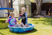 Load image into Gallery viewer, Two 11 year old girls sitting in a blue paddling pool in a garden. They are holding a red and a blue EcoSplat Reusable Water Balloon ready to throw them and are laughing. There are red water balloons lying on the grass.
